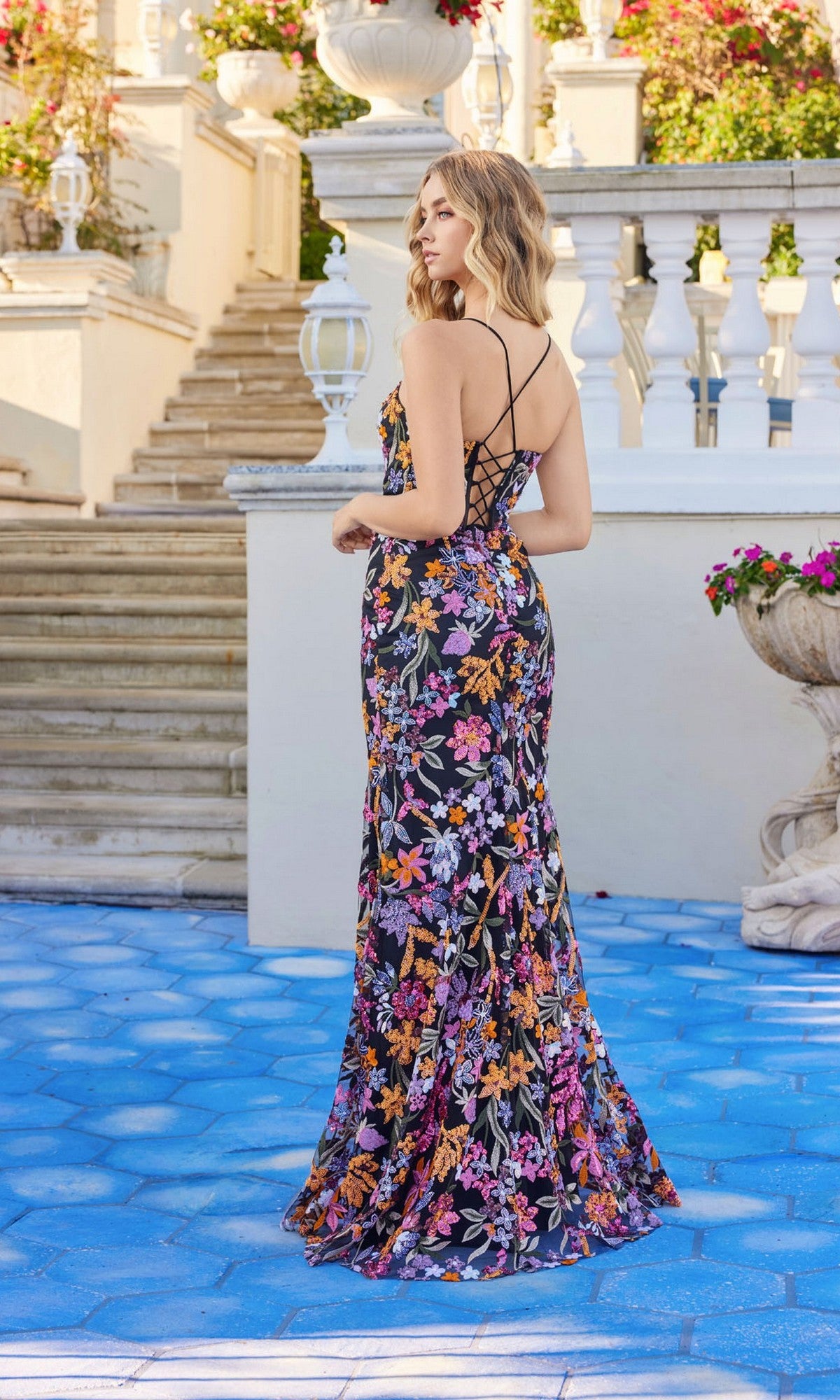 A woman wears the Juliet JT2498A Long Floral-Sequin Prom Dress, a backless, floor-length gown with a sweetheart neckline, as she stands on a blue tiled patio near white railings amid flowers and greenery.